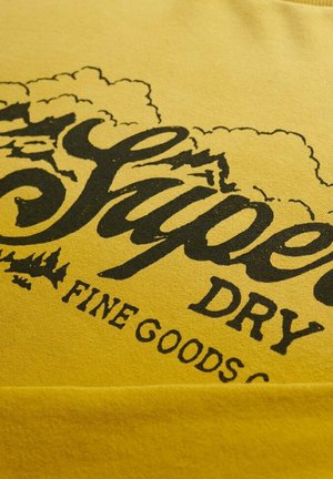 Gul stof med sort stiliseret tekst, der læser "Superdry Fine Goods Co" og et grafisk billede af skyer og træer.