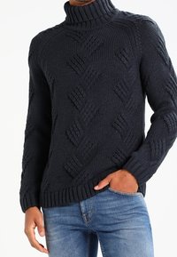 Fetter gestrickter navyfarbener Rollkragenpullover mit strukturiertem Rautenmuster, gerippten Bündchen und Saum. Kombiniert mit hellblauen Jeans.
