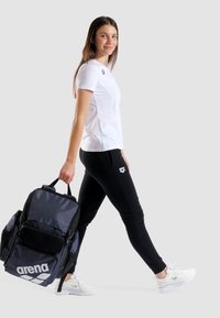  Sorte træningsbukser og en hvid t-shirt, med en stor mørkeblå arena rygsæk med en netlomme. Hvide sneakers.
