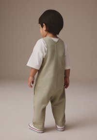 Gaiši zaļa kokvilnas kombinezons bez piedurknēm ar aizmugurējo kakla izgriezuma detaļu, combinēts ar baltu kreklu un sarkanām apaviem ar akcents.