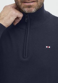 Marineblå zip-up genser med høy krage, ribbestruktur og en liten brodert logo med røde, hvite og blå striper på brystet.