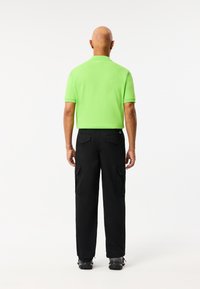 Neon groene polo-shirt met korte mouwen, gecombineerd met zwarte cargobroek met zijzakken. Het uitzicht is van achter.
