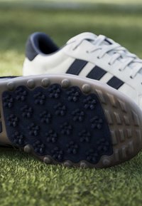 Λευκό και ναυτικό αθλητικό παπούτσι με τάπες στη σόλα που ακουμπάει στο γρασίδι, δείχνοντας κοντινή εικόνα της υφής της πρόσφυσης και τρεις ναυτικές ρίγες στο πλάι.