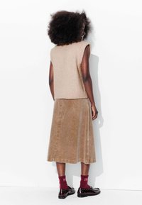 Un chandail sans manches en tricot beige associé à une jupe midi en velours côtelé brun. Le mannequin porte des chaussettes bordeaux et des chaussures noires, tourné dos au spectateur.