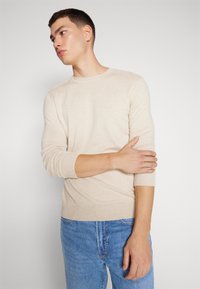 Pulôver de malha bege com gola redonda e punhos ajustados, combinado com jeans azul claro. Textura suave e design de ajuste padrão.