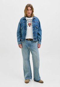 Noor inimene pikkade juustega, kes kannab teksajakki, valget graafilise kujundusega "Hellfire Club" T-särki, heledaid siniseid laiadest pükstest ja beeže saapaid.