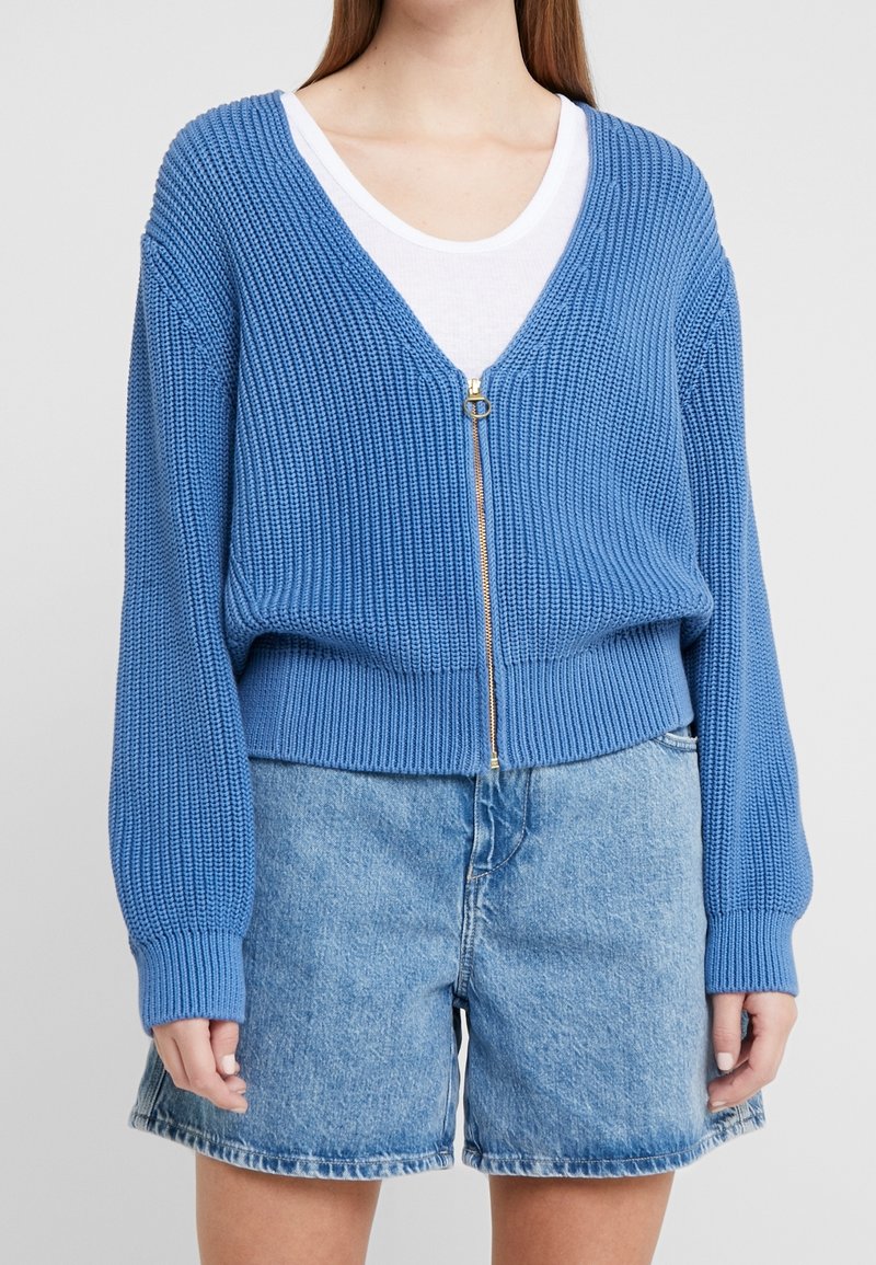 Blå stickad cardigan med V-ringning och guldblixtlås, med ribbstickade ärmslut och nederkant. Kommer med ljusblå denimshorts.