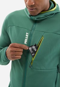 Green fleece jacket with a zippered chest pocket. Yellow zipper accent and a visible label for protein contents being retrieved from the pocket.