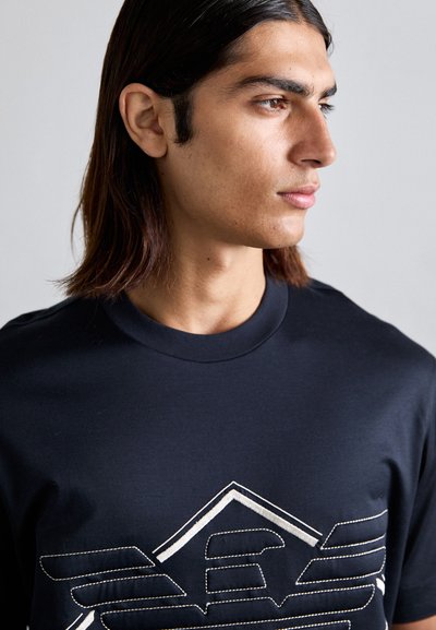 Navy blauwe katoenen t-shirt met een wit gestikt geometrisch ontwerp op de voorkant en een ronde halslijn. Soepele textuur, korte mouwen.