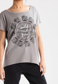 Camiseta gris de corte holgado, cuello redondo y mangas cortas. Presenta un gráfico de mandala negro con el texto "salvaje y libre" en el centro.