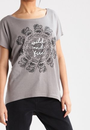 T-shirt grigio con vestibilità ampia, scollatura rotonda e maniche corte. Presenta una grafica nera di mandala con il testo "selvaggio e libero" al centro.