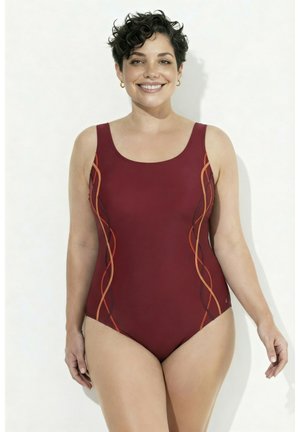 Femme souriante aux cheveux courts et bouclés, portant un maillot de bain une pièce bordeaux avec des rayures latérales ondulées orange et noires, debout sur un fond blanc.