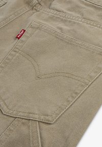 Μπεζ τζιν ύφασμα με υφή, που διαθέτει έντονο σχέδιο πίσω τσέπης και κόκκινη ετικέτα μάρκας κοντά στη ραφή.