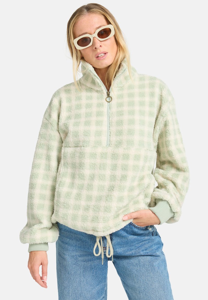A woman poses confidently, wearing a light green checkered fleece jacket and sunglasses, with hands on her hips and a relaxed expression.