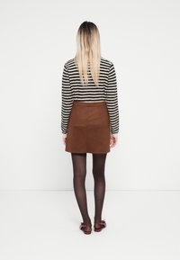 Brauer Wildlederrock mit tailliertem Bund, kombiniert mit einem schwarz-weiß gestreiften Strickpullover und dunklen semitransparenten Strumpfhosen. Rote Sandalen sind sichtbar.