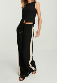 Zwarte geribbelde crop top met knoopsluiting, gecombineerd met losse zwarte broek met beige zijstreep en een verstelbare trekkoord taille.