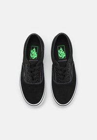 Baskets en toile noires avec coutures blanches et semelles en caoutchouc. Logo vert sur la semelle intérieure, design à bout rond, style classique à lacets.