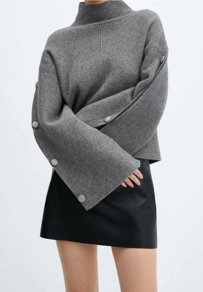 Personne portant un pull gris surdimensionné en maille côtelée avec des manches larges et des boutons le long des bras, associé à une mini-jupe noire.