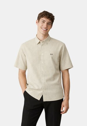 Giovane uomo sorridente che indossa una camicia beige a maniche corte con sottili righe e pantaloni neri, in piedi con una mano in tasca.