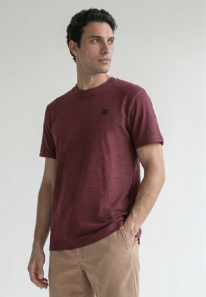 Homme aux cheveux bouclés foncés portant un t-shirt bordeaux à manches courtes et un pantalon beige en velours côtelé, regardant vers sa gauche avec une main dans la poche.