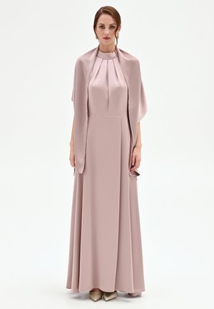 Femme en robe longue en satin rose poudré avec un corsage plissé et un châle assorti, posant devant un fond blanc uni.