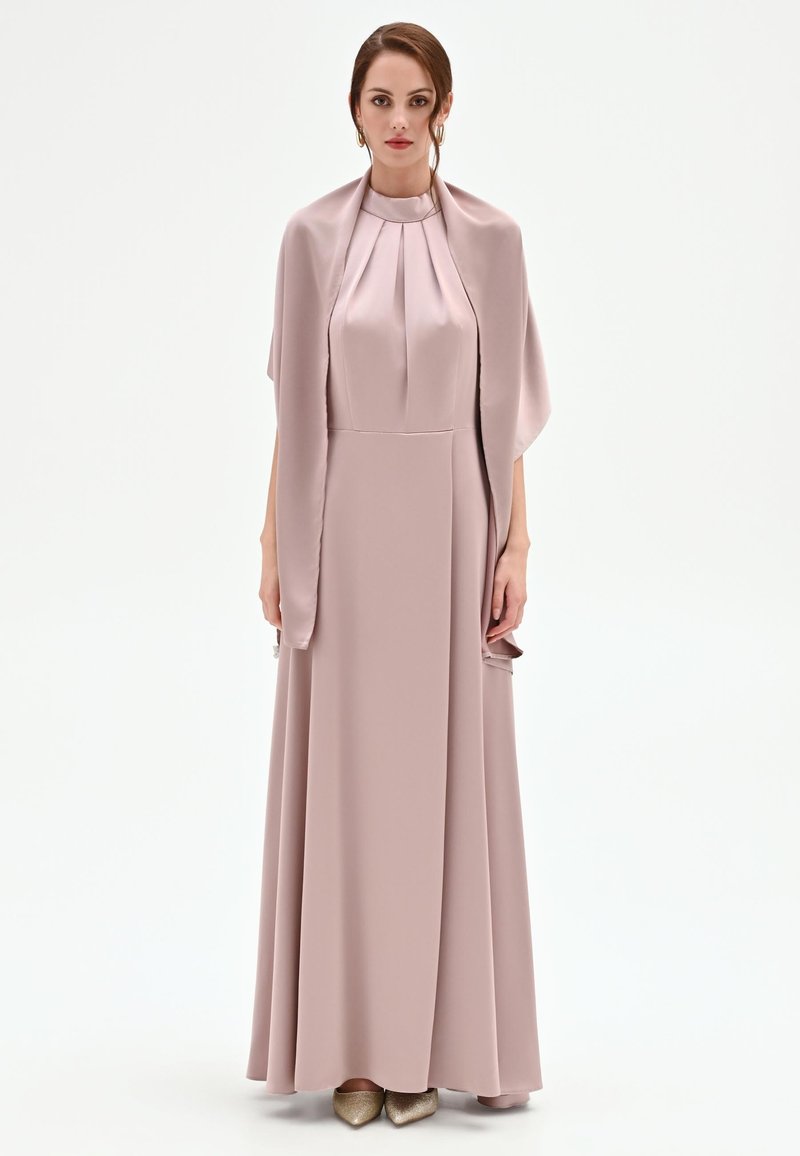 Femme en robe longue en satin rose poudré avec un corsage plissé et un châle assorti, posant devant un fond blanc uni.