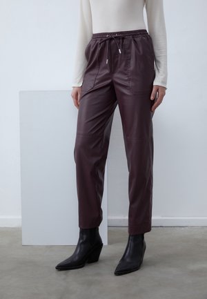 Personne portant un pantalon en cuir marron foncé avec taille à cordon et des bottines noires à bout pointu, debout sur un sol gris sur fond blanc.