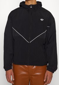Černá kapucňová bunda se stříbrnými zipy, se bílými diagonálními pruhy a texturovanou látkou. Logo Adidas na levé hrudi.