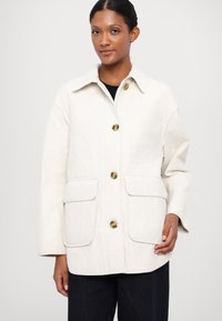 Light beige quilted jacket with a collar, front button closure, and two large front pockets. Features a textured fabric and subtle sheen.
