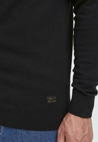 Schwarzer Strickpullover mit geripptem Saum und Bündchen, mit einem kleinen braunen Etikett, auf dem "ISOLID Since 1999" steht, unten links.