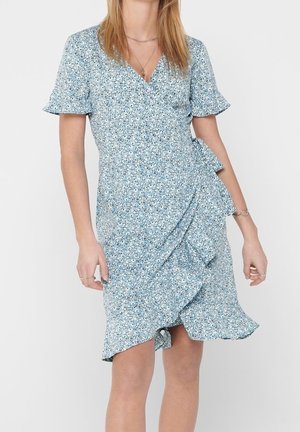 Vrouw draagt een lichtblauwe bloemenprint wikkeljurk met korte fladdermouwen en een gerimpelde zoom, staand tegen een effen achtergrond.