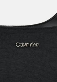 Crni materijal s uzorkom u reljefu, na kojem je istaknuto srebrno Calvin Klein logo.