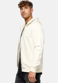 Crème kleur zip-up hoodie met een gevoerde capuchon, voorzakken en ribgebreide manchetten. Heeft een logo-detail op de capuchon en een ontspannen pasvorm ontwerp.