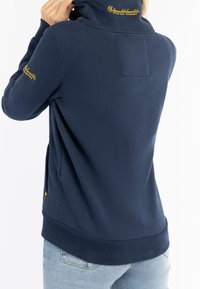 Pullover blu navy con colletto alto, caratterizzato da ricami dorati sulla spalla e una texture liscia. Tasche laterali visibili.