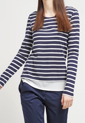 Femme portant un t-shirt à manches longues rayé horizontalement bleu marine et blanc par-dessus un haut blanc et un pantalon bleu marine, debout devant un fond uni.