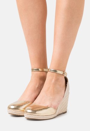 Pieds portant des chaussures compensées dorées métalliques avec des brides à la cheville et des semelles plates à texture tissée sur un fond clair uni.