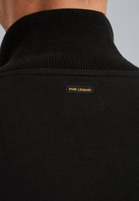 Mustapusero, jossa on korkea ribbireuna, ja takana on pieni suorakulmainen etiketti, jossa lukee "PME LEGEND" keltaisella tekstillä.