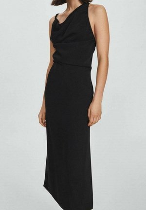 Femme portant une robe noire asymétrique sans manches avec un décolleté drapé et un ourlet à la cheville, debout devant un fond clair uni.