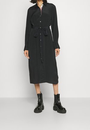 Mujer con vestido midi negro de manga larga, abotonado, con cinturón en la cintura y botas negras gruesas hasta el tobillo, de pie sobre un suelo gris.