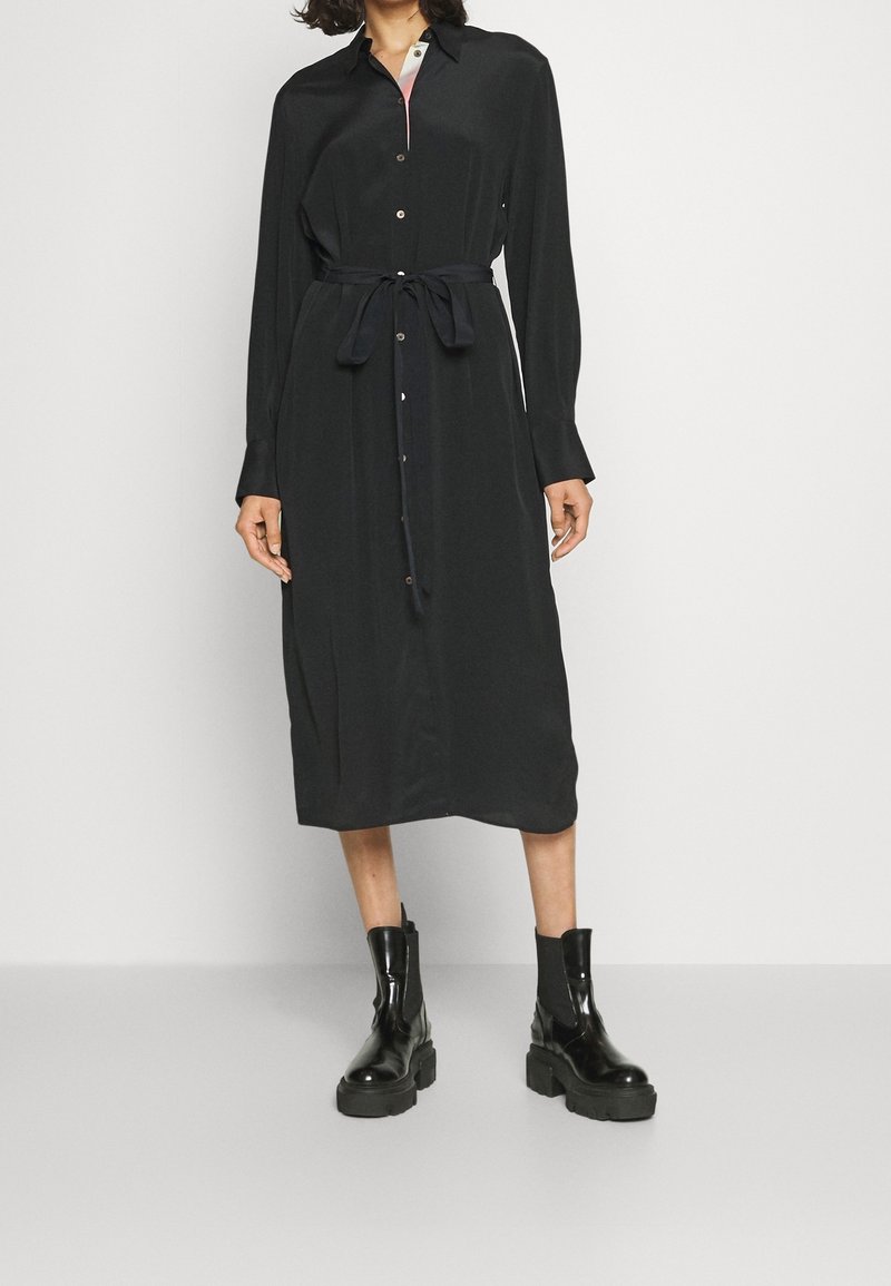 Femme portant une robe midi noire à manches longues boutonnée avec une ceinture à la taille et des bottines noires épaisses, debout sur un sol gris.
