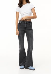 Kvinde iført en hvid cropped t-shirt, højtaljede mørkegrå flared jeans og lyseblå sneakers, stående foran en hvid baggrund.