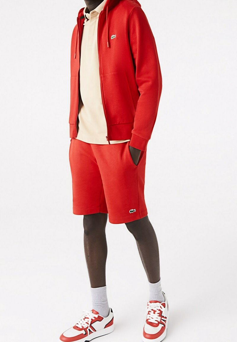 Homme portant un sweat à capuche rouge zippé et un short assorti avec des baskets blanches et rouges, chemise beige en dessous, debout les mains dans les poches.