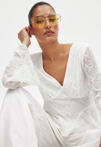 Weiße bestickte Bluse mit V-Ausschnitt und Glockenärmel, kombiniert mit gelb getönten Sonnenbrillen. Weiche, florale Textur und gestuftes Design.