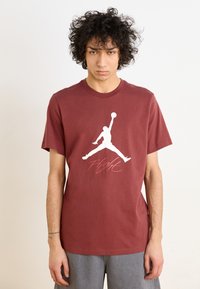 Mørkerød bomulds t-shirt med et hvidt Jordan-logo og ordet "Flight" i en kursiv pink skrifttype. Korte ærmer og rund hals.