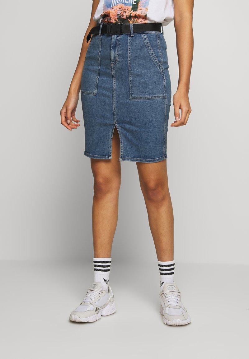Femme portant une jupe en denim bleu avec une fente à l'avant, taille ceinturée, baskets blanches et chaussettes blanches à rayures noires, debout devant un fond uni.