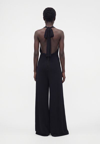 Femme debout de dos portant une combinaison noire à col halter, dos ouvert et pantalon à jambes larges sur fond blanc.