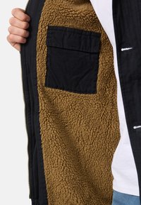 Veste noire zippée avec une doublure intérieure en fausse fourrure marron. Comprend une poche intérieure en tissu noir. La doublure texturée apporte chaleur et contraste.