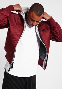Giacca bomber rossa in nylon con fodera a cappuccio grigia, polsini a coste e chiusura frontale con zip; presenta una patch sul petto e tasche laterali.