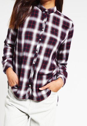 Kobieta w granatowo-biało-czerwonej koszuli w kratę zapinanej na guziki z długimi rękawami, ręce w kieszeniach, w połączeniu z jasnymi spodniami.