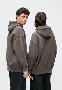 Γκρι φούτερ με κουκούλα και άνετη εφαρμογή, με φαρδιά μανίκια, ριμπ τελειώματα και τσέπη καγκουρό, φορεμένο με σκούρο τζιν.